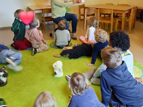 Kinder sitzen und liegen im Kreis, Erzieherin hält ein Buch mit Wolfsbildern hoch