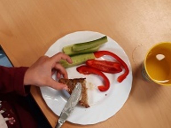 Jemand macht sich eine Scheibe Brot, auf dem Teller liegen Paprikastreifen und Gurkenscheiben