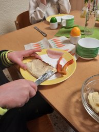 Blick auf einen Frühstücksteller, Kinderhände schmieren Butter auf eine Scheibe Brot, Schinkenwurst und Paprikastreifen liegen danebenika und