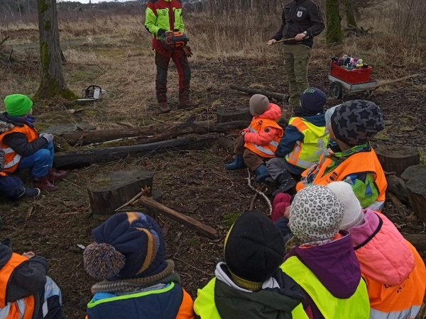 Kinder sitzen im Kreis auf dem Waldboden. Eine Försterin und ein Forstwirt zeigen ihr Werkzeug.