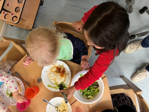 Blick von oben auf einen Esstisch, Kind füllt kleinerem Kind Salat auf