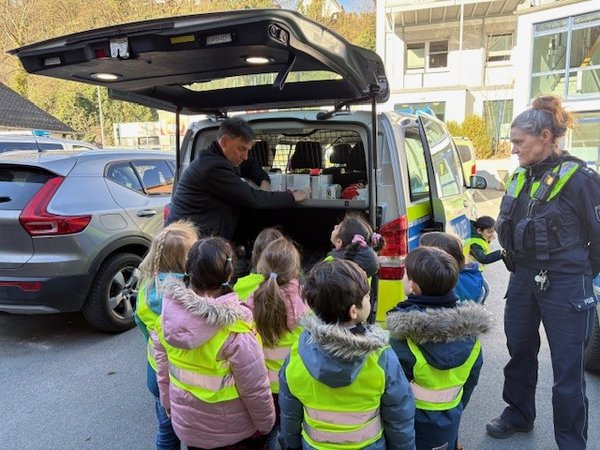 Kinder und 2 Polizeibedienstete stehen an der geöffneten Heckklappe eines Polizeifahrzeugs
