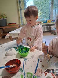 Ein Junge im Malkittel pinselt ein Vogelhäuschen grün an