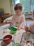 Ein Junge im Malkittel pinselt ein Vogelhäuschen grün an