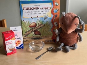 Eine Plüschameise sitzt neben einem Buch und Packungen mit Zucker und Salz auf einem kleinen Tisch
