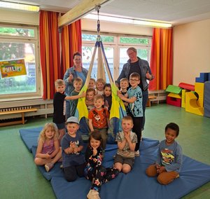 Gruppenfoto von Frau Schulze, Frau Krueger und KiTa-Kindern, die in und auf Turnmatten unter der neuen Schaukel sitzen