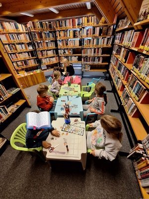 Kinder sitzen an Tischen in der Bücherei, eine Mitarbeiterin sitzt vor Kopf und liest eine Geschichte vor.